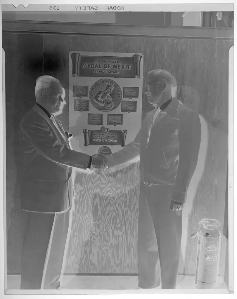 Untitled (two men shaking hands in front of "medal of merit" service award) by Jack Rodden Studio