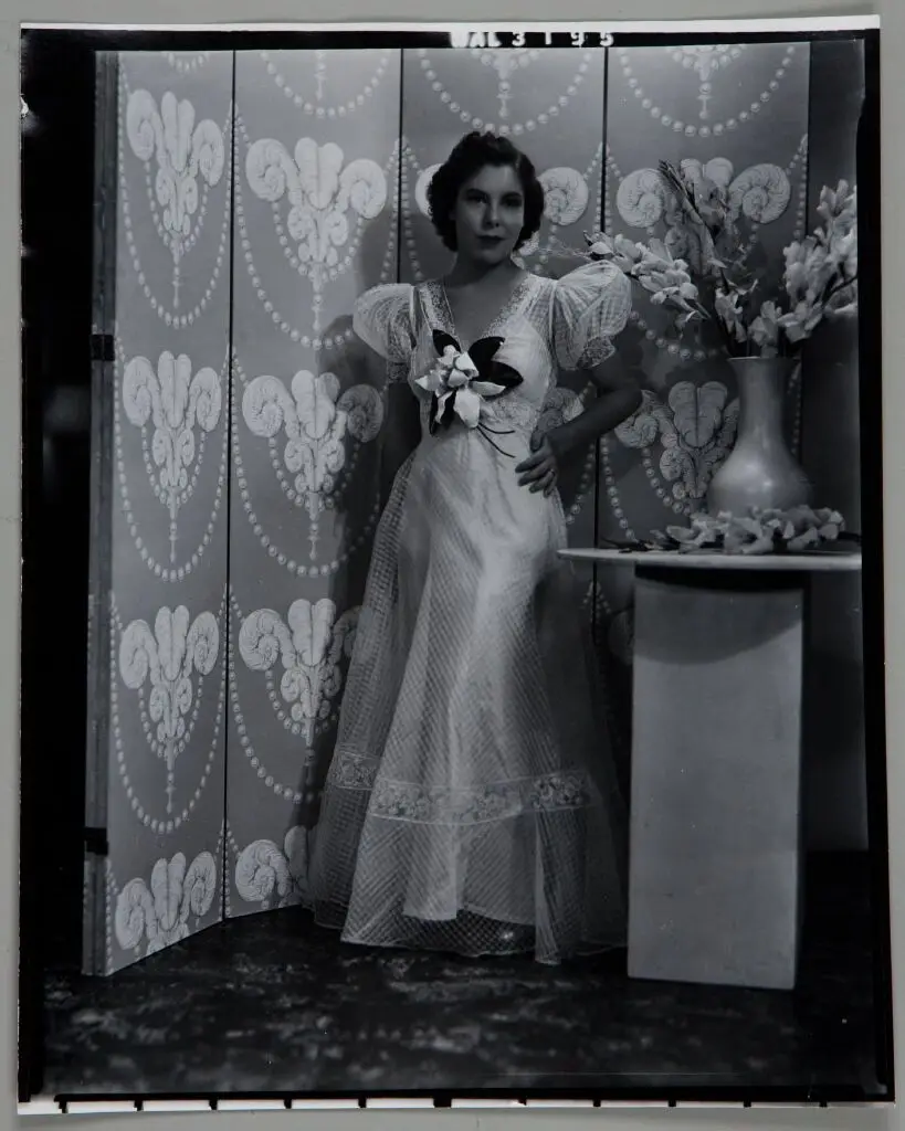Untitled (full length portrait of woman in dress with flower at chest, by ornate screen and vase of flowers) by Paul Gittings