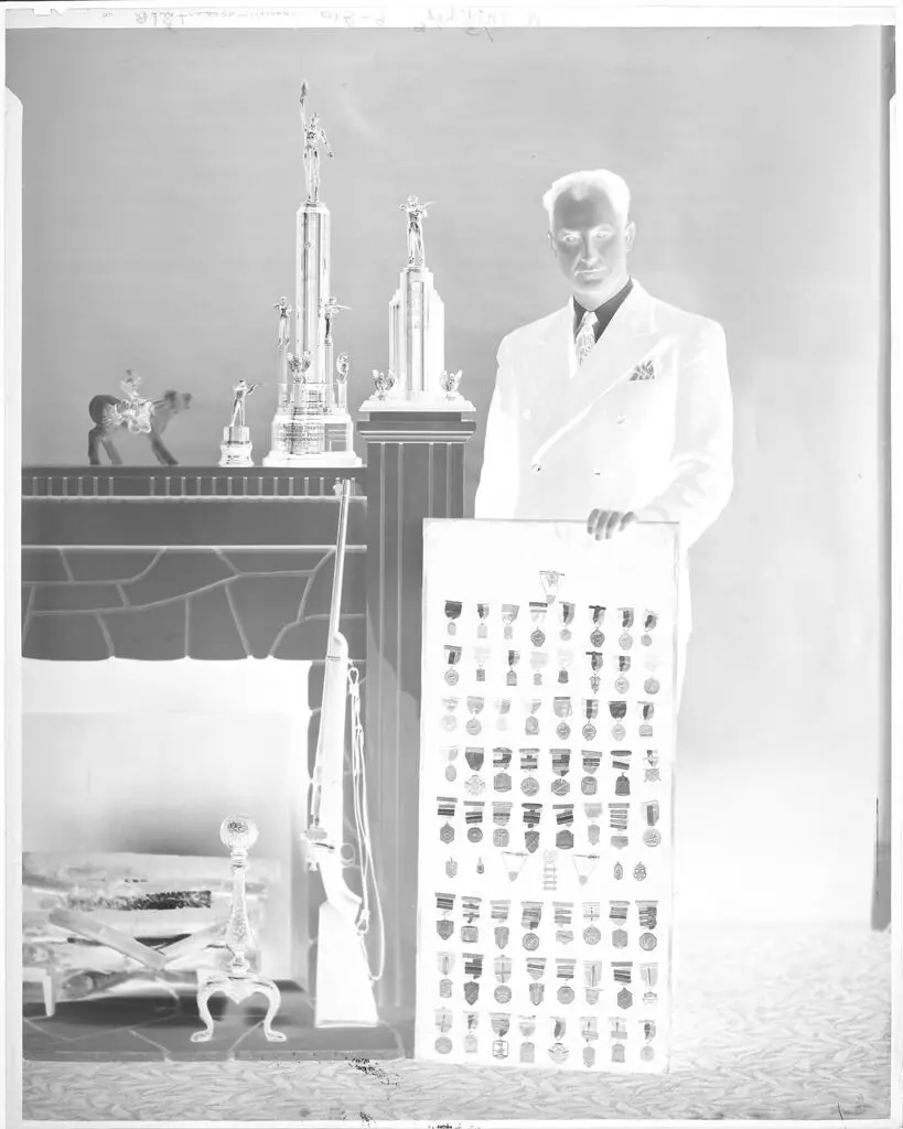 Untitled (man posing with medals and trophies for shooting with his rifle by fireplace) by John Deusing